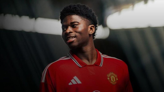 Young man in a red Manchester United jersey smiling indoors, facing left.