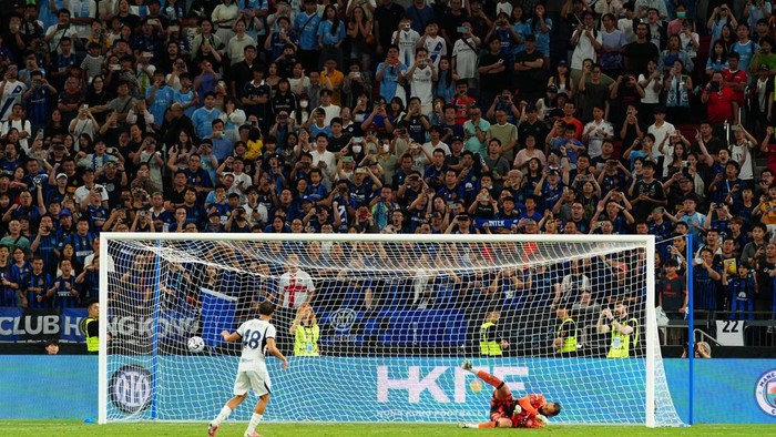 Soccer goal action: a white-uniformed player near the net as the goalkeeper sprawls on the ground; packed stadium in the background.