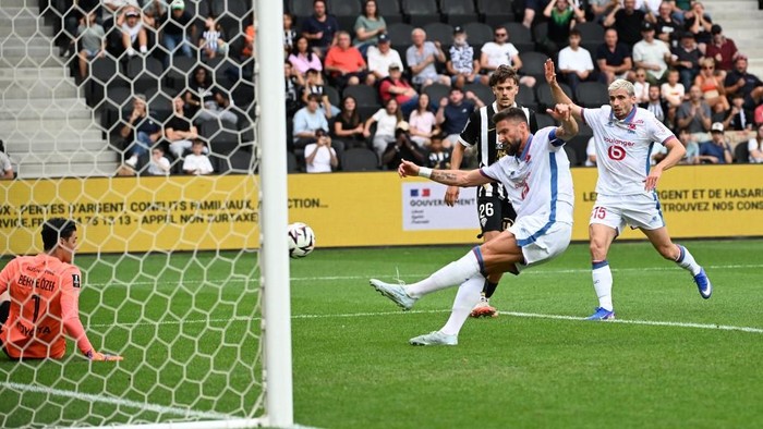 Soccer scene with a goalkeeper in orange on the left near the goal, a white-clad player kicking the ball, and another defender nearby as spectators watch from the stands.