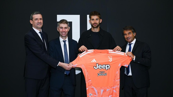 Four men in suits present an orange Juventus jersey against a dark wall backdrop.