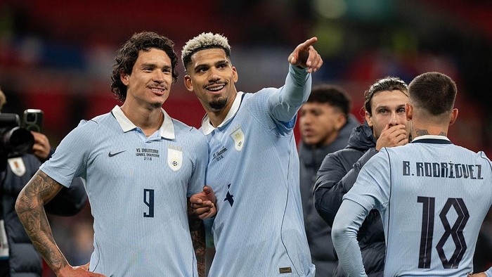 Three football players in light‑blue jerseys celebrate on the field; one points forward as another smiles, with a teammate seen from the back wearing number 18.