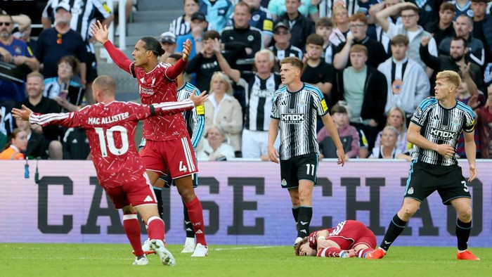 Soccer match moment: players in red celebrate a goal with arms raised as a teammate in red wears number 10 (Mac Allister) nearby; black-and-white striped players stand by, while a red player lies on the grass.