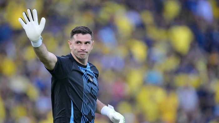 Male soccer goalkeeper raises gloved hand in a wave to the crowd during a match, dark jersey with blue accents, yellow-tinged stands in background