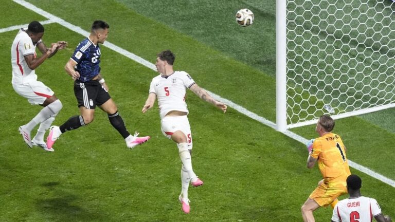 Football scramble near the goal: attackers contesting a ball in the air as the goalkeeper in orange watches from the right edge of the goal line.