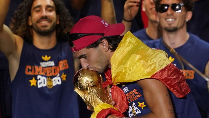Athlete kissing a gold trophy while draped in a red and yellow flag, teammates celebrate in the background.