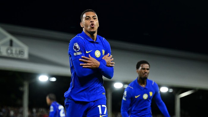 Two soccer players in blue celebrate a goal with crossed arms on the field at night, stadium lights behind them.