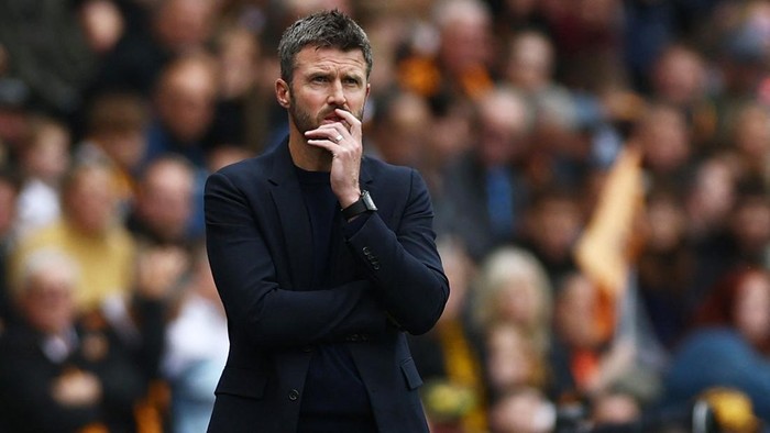 Football manager on the sideline, arms crossed with a hand to the chin, deep in thought as the crowd watches the match.