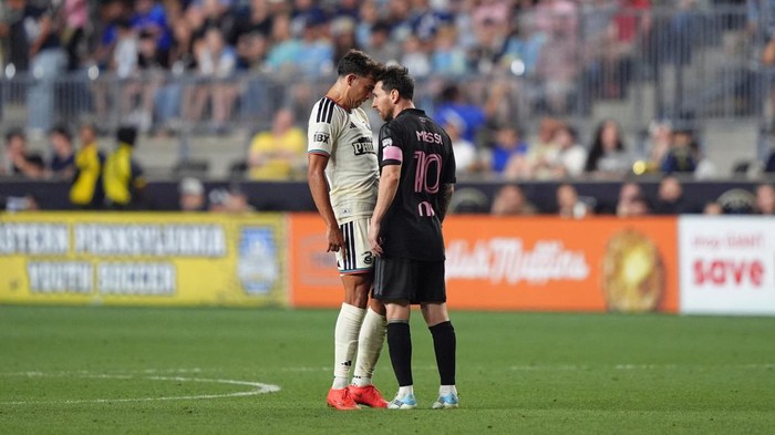 Two soccer players in contrasting kits stand nose-to-nose on a green field amid a cheering crowd in the stands.
