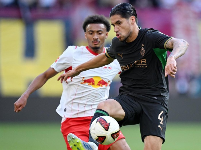 Two soccer players contest the ball mid‑play; the player in black controls the ball while the opponent in white and red closes in on a grassy field.