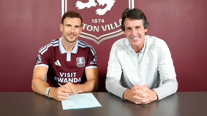 Two men sit at a table signing a document, with an Aston Villa backdrop behind them.