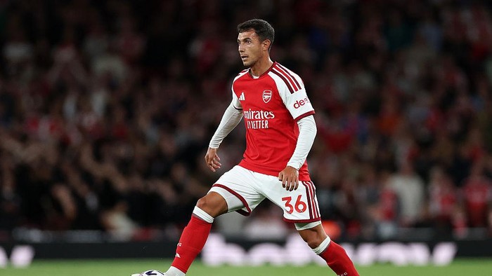 Arsenal player in red and white kit dribbling with the ball during a match, crowd blurred in the background.