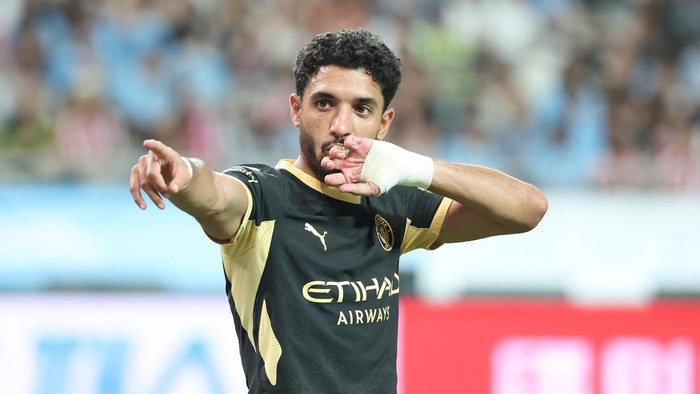 Soccer player in a black-and-gold jersey gestures with his hand near his lips during a celebration, with a blurred stadium crowd in the background.