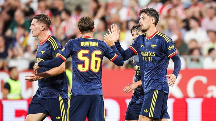 Soccer players in blue away kits celebrate on the pitch, high-fiving after a play; the back of a player wearing number 56 is visible.