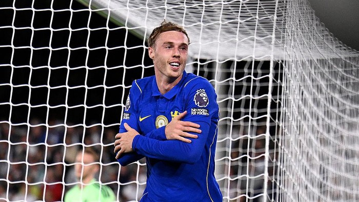 Soccer player in a blue kit celebrates in front of the goal, arms crossed with the net behind him.