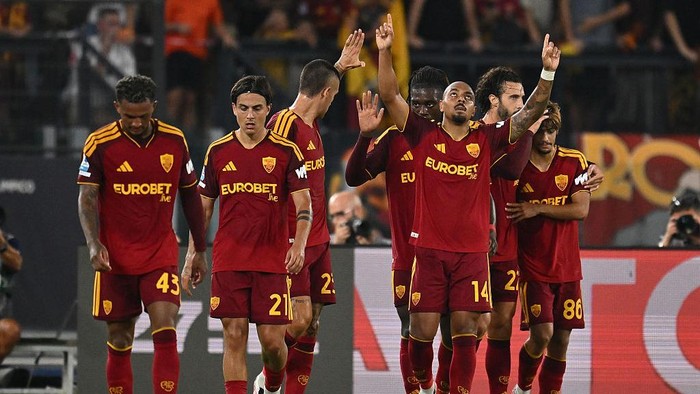 AS Roma players in red and yellow kits celebrate on the pitch, some raising their hands in celebration.
