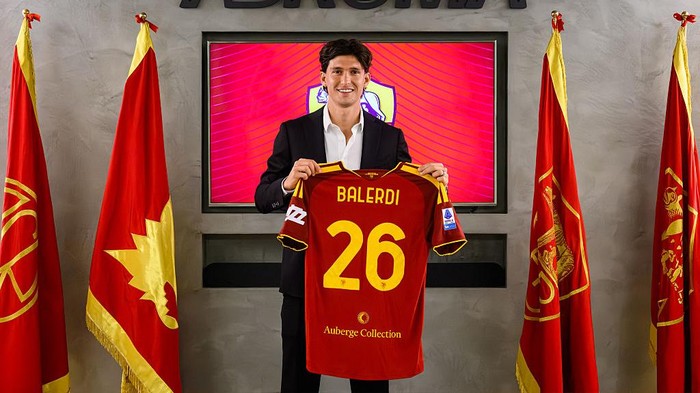 Man in a suit holding a red soccer jersey with the name BALERDI and number 26, flanked by red and gold flags in a formal room