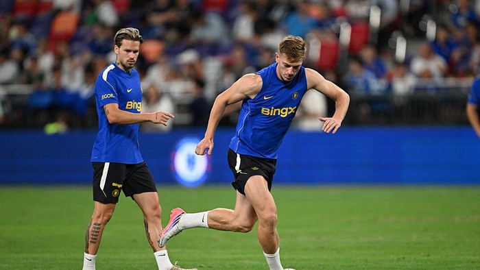 Two male soccer players in blue training shirts on a green field, one kicking the ball during practice on a stadium pitch.