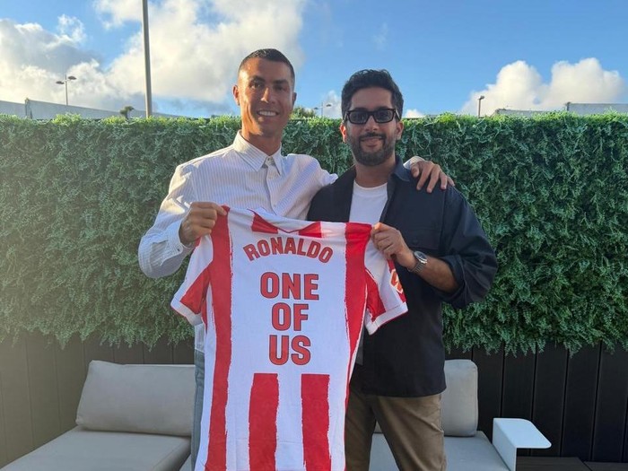 Two men pose outdoors, holding a red-and-white striped soccer jersey that reads 'RONALDO ONE OF US'.