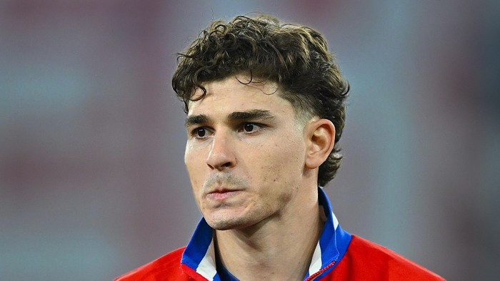 Close-up of a young man with short dark curly hair wearing a red sports jacket with a blue collar, looking focused on something off-camera.