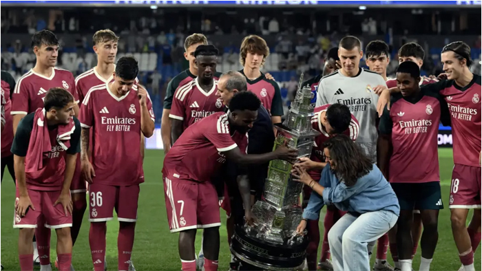 Real Madrid players in pink away kits celebrate on the pitch around a large trophy, lifting it in triumph after a victory.
