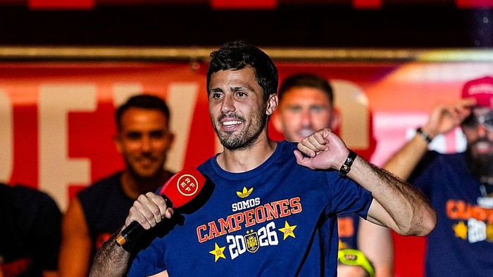 Man in a blue Campeones 2026 shirt speaks into a microphone at a celebration stage, with his fist raised in victory as teammates smile in the background.