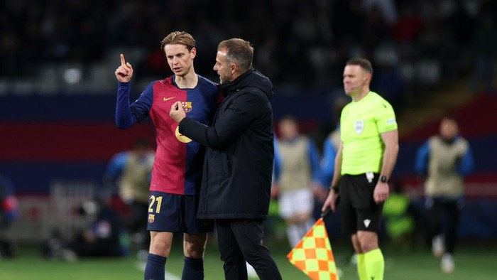 Football coach in a dark coat talks to a Barça player in red-blue kit on the field, with a referee in neon nearby.