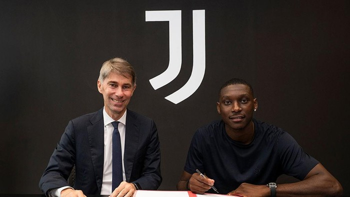 Two men sit at a table signing a document, with a large Juventus logo behind them.