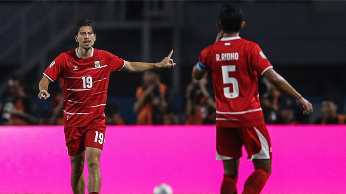 Two soccer players in red jerseys celebrate on the field; player number 19 points with his arm extended as teammate number 5 stands nearby in front of a pink advertising board.