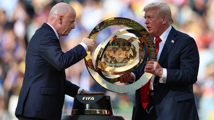Two men in suits hold a large gold FIFA World Cup trophy during a ceremony, with a blurred crowd in the background.