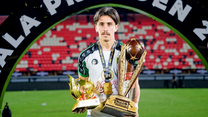 Male soccer player standing on a pitch, holding two trophies and a gold medal; stadium stands behind him under a curved arch.