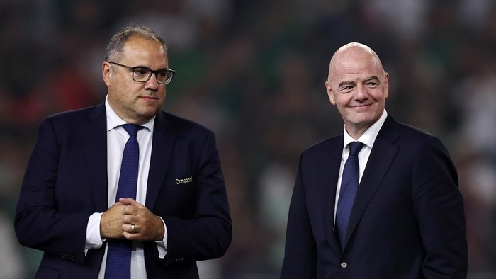 Two men in dark suits stand on a blurred stadium background, one wearing glasses and a tie, the other bald and smiling slightly.