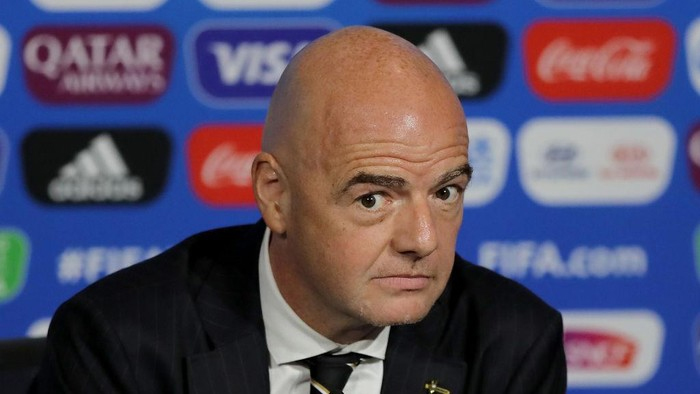 Bald man in a dark suit and striped tie at a press conference against a blue sponsor backdrop, looking toward the camera.