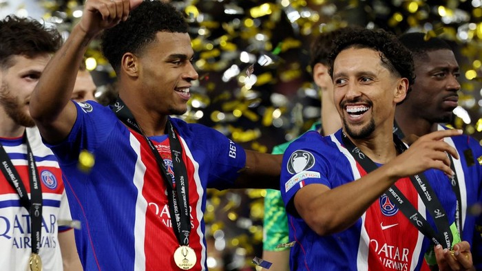 Two Paris Saint-Germain players celebrate victory, wearing blue jerseys and gold medals as confetti falls.