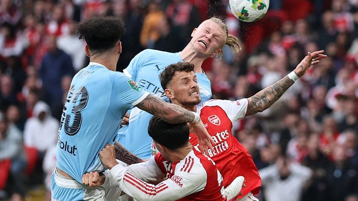 Players in blue and red collide in midair, leaping to head the ball during a crowded match scene.”,