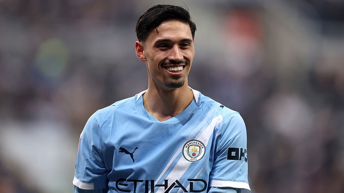 Male soccer player in Manchester City light-blue kit smiling on the pitch.