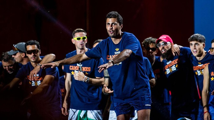 A group of men in blue shirts, led by a man in front gesturing as if directing or dancing, during a team celebration.