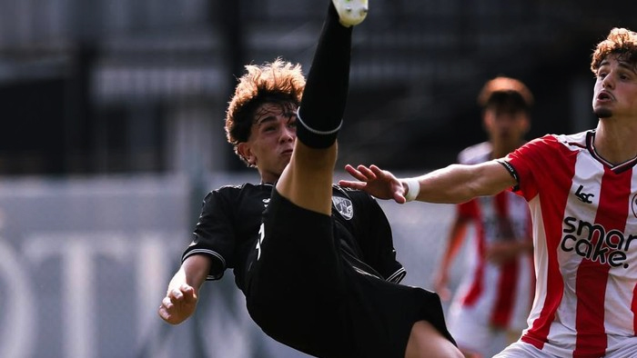 Soccer players in a high kick challenge during a match; one player in black raises a leg while an opponent in red-and-white stripes reaches forward.