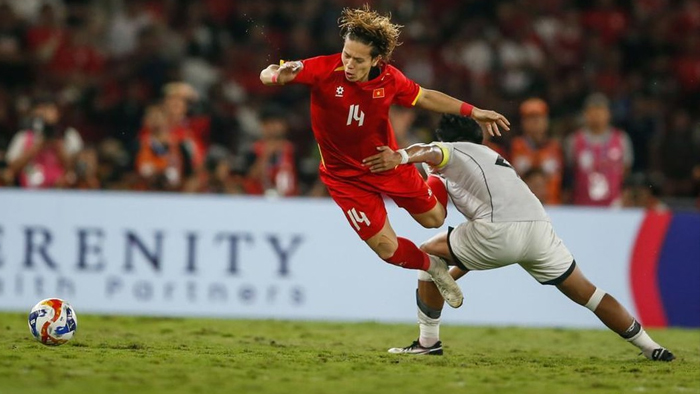 Soccer player in red jersey #14 dribbles the ball while an opposing player in white challenges from behind on a grassy field with a cheering crowd in the background.