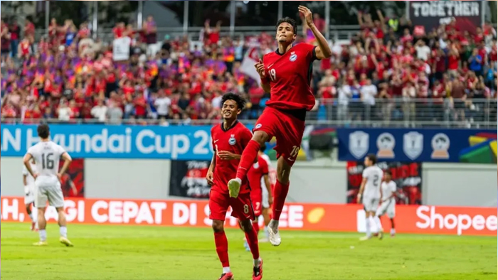 Two players in red jerseys celebrate on a soccer pitch as one jumps mid‑air, with cheering fans in red in the stands.