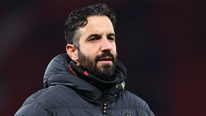 Man with a dark beard and short, wavy hair wearing a navy puffer jacket outdoors, looking off to the side with a serious expression.