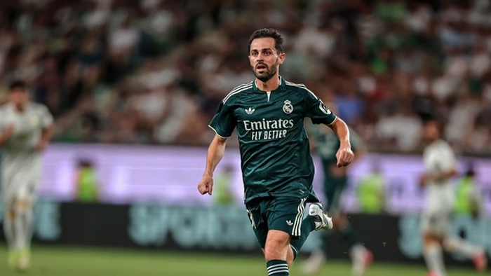 Real Madrid player in a green away kit runs on the pitch during a match with a blurred crowd behind him.
