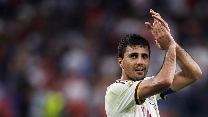 Soccer player in a white jersey clapping with hands raised, stadium crowd blurred in the background.