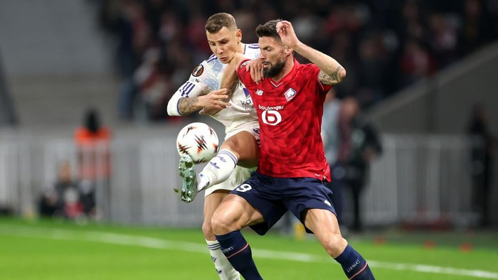 Two soccer players contest the ball in a forward duel on a green pitch; one in white kit, another in red.