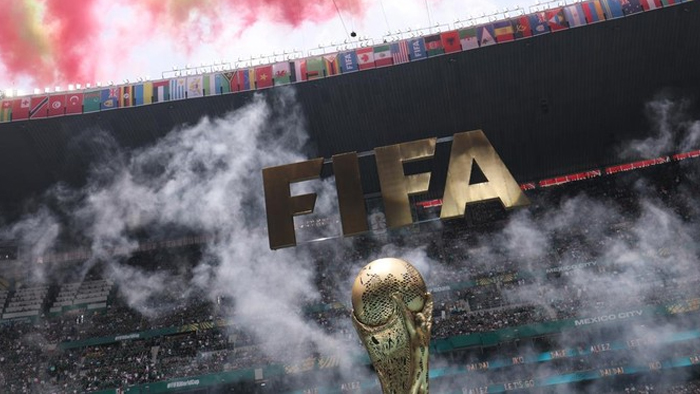 Golden FIFA World Cup trophy on the field with dramatic red smoke and flag-filled stands in the background.