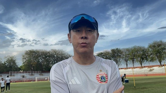 Athlete in a light gray jersey with crest stands on a soccer field, wearing a blue cap and sunglasses.