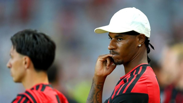 Male soccer player in a white cap and red-and-black jersey, touching his lips, on the sidelines during a match.