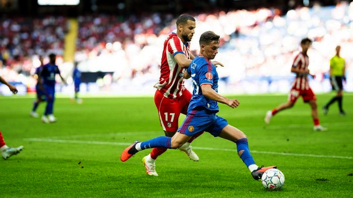 Soccer players in blue and red striped kits chase the ball on a green field during a match.