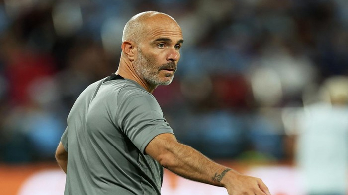 Bald man with a gray beard in a gray athletic shirt on a stadium field, turning to look over his shoulder.