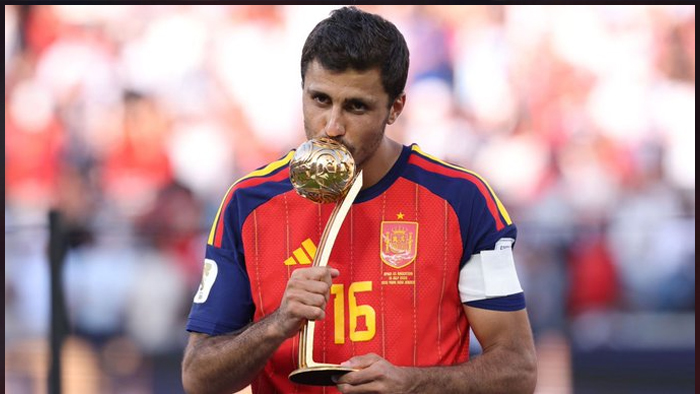 Male footballer in a red jersey kisses a gold trophy on the field with a blurred crowd in the background.
