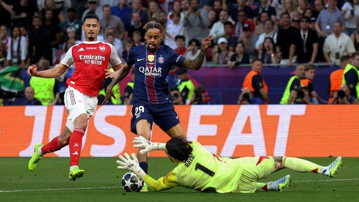 PSG player in blue dribbles toward goal as Arsenal defender and goalkeeper dive to stop the shot on a packed stadium field.